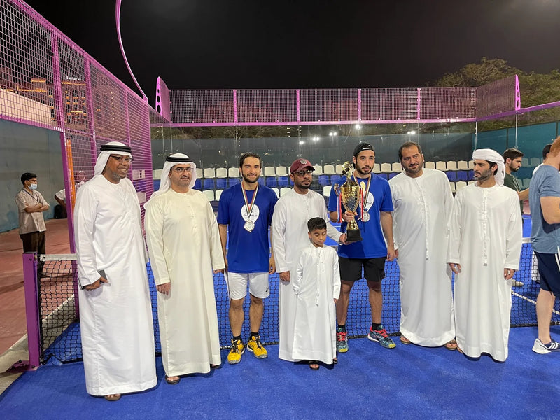 Group of people, including men in traditional Middle Eastern attire and a child, standing on a padel court with a trophy.